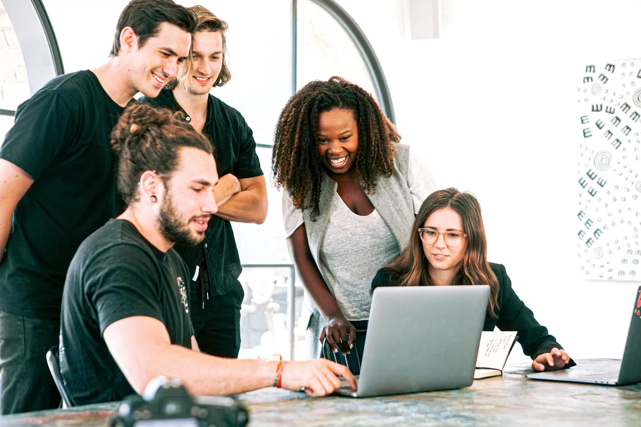 Group of five people around a work table looking at a laptop screen in a bright office.